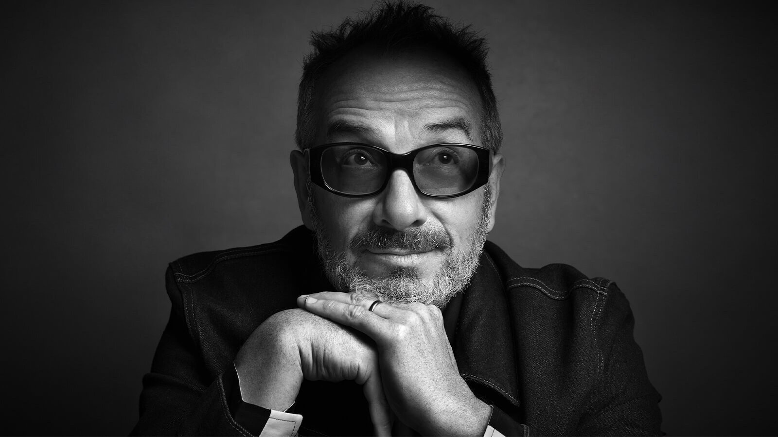 A black and white portrait of singer, songwriter, and producer Elvis Costello wearing dark-tinted glasses and a wedding band.