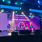 A large screen reads "5th Annual Songwriters Music Workshop" against a purple backdrop. The screen sits atop a brightly lit, empty stage.